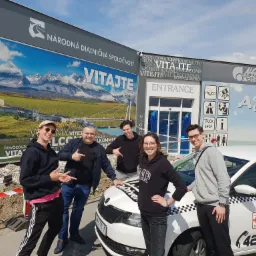 Grupa pięciu osób pozuje przy białej taksówce z napisem 'Taxi Łódź' na tle budynku z napisami powitalnymi w różnych językach i billboardem z górskim krajobrazem.