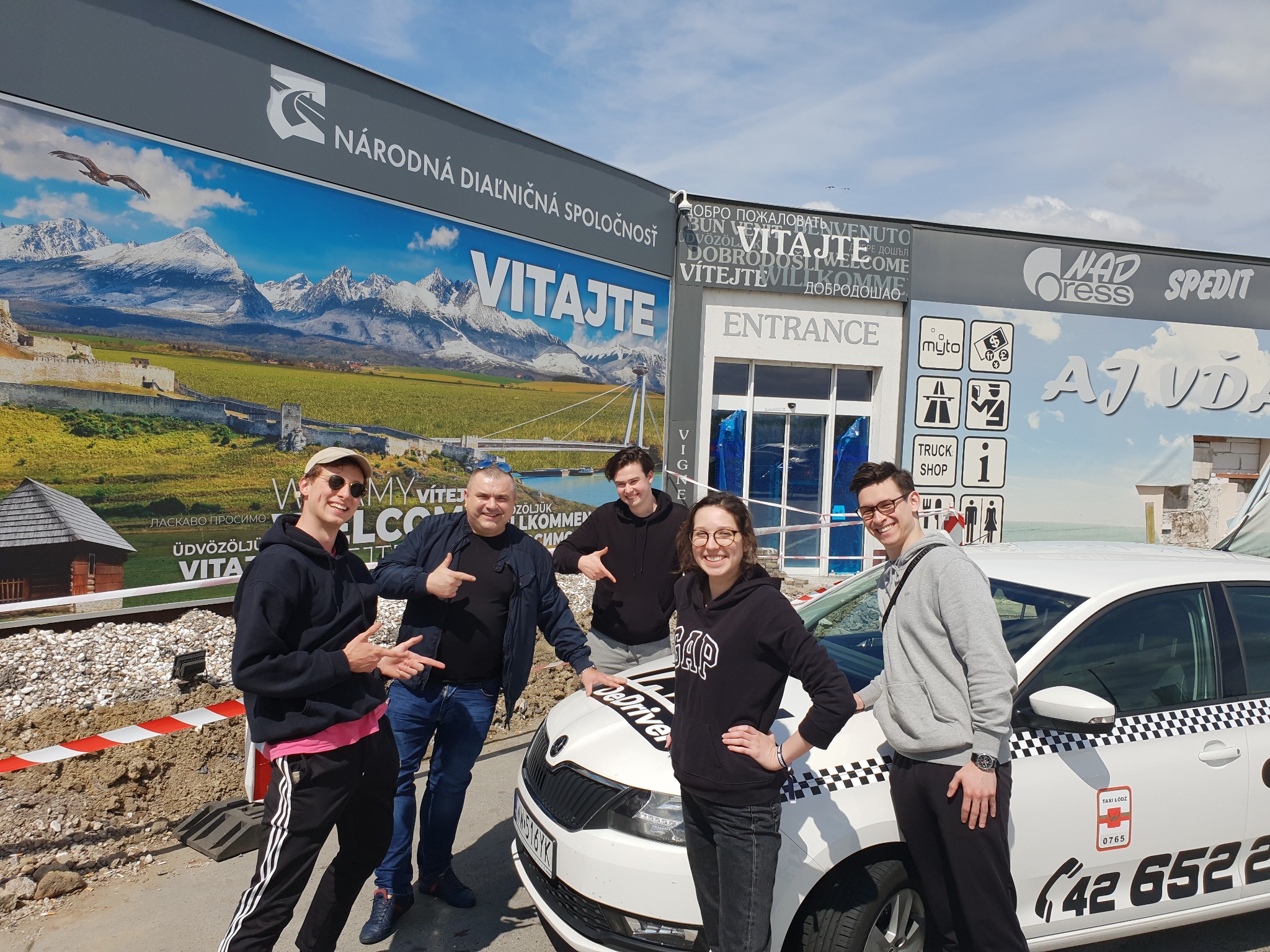 Grupa pięciu osób pozuje przy białej taksówce z napisem 'Taxi Łódź' na tle budynku z napisami powitalnymi w różnych językach i billboardem z górskim krajobrazem.