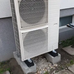 Biała pompa ciepła Panasonic na betonowych blokach, z drobnym żwirem wokół podstawy. Widoczne okno w tle i betonowa kostka brukowa.