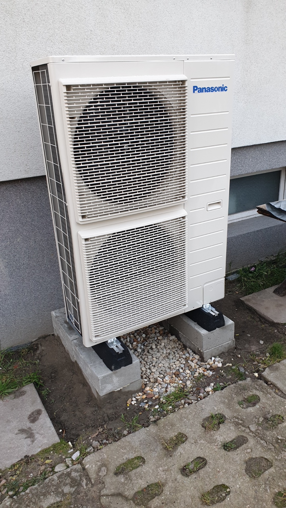 Biała pompa ciepła Panasonic na betonowych blokach, z drobnym żwirem wokół podstawy. Widoczne okno w tle i betonowa kostka brukowa.