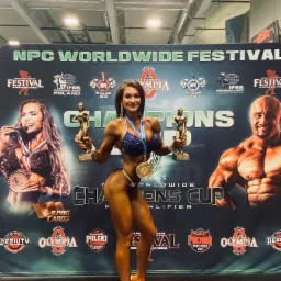 Kobieta w bikini pozuje z dwoma statuetkami i medalami na tle baneru NPC Worldwide Festival, prezentując wyrzeźbioną sylwetkę.