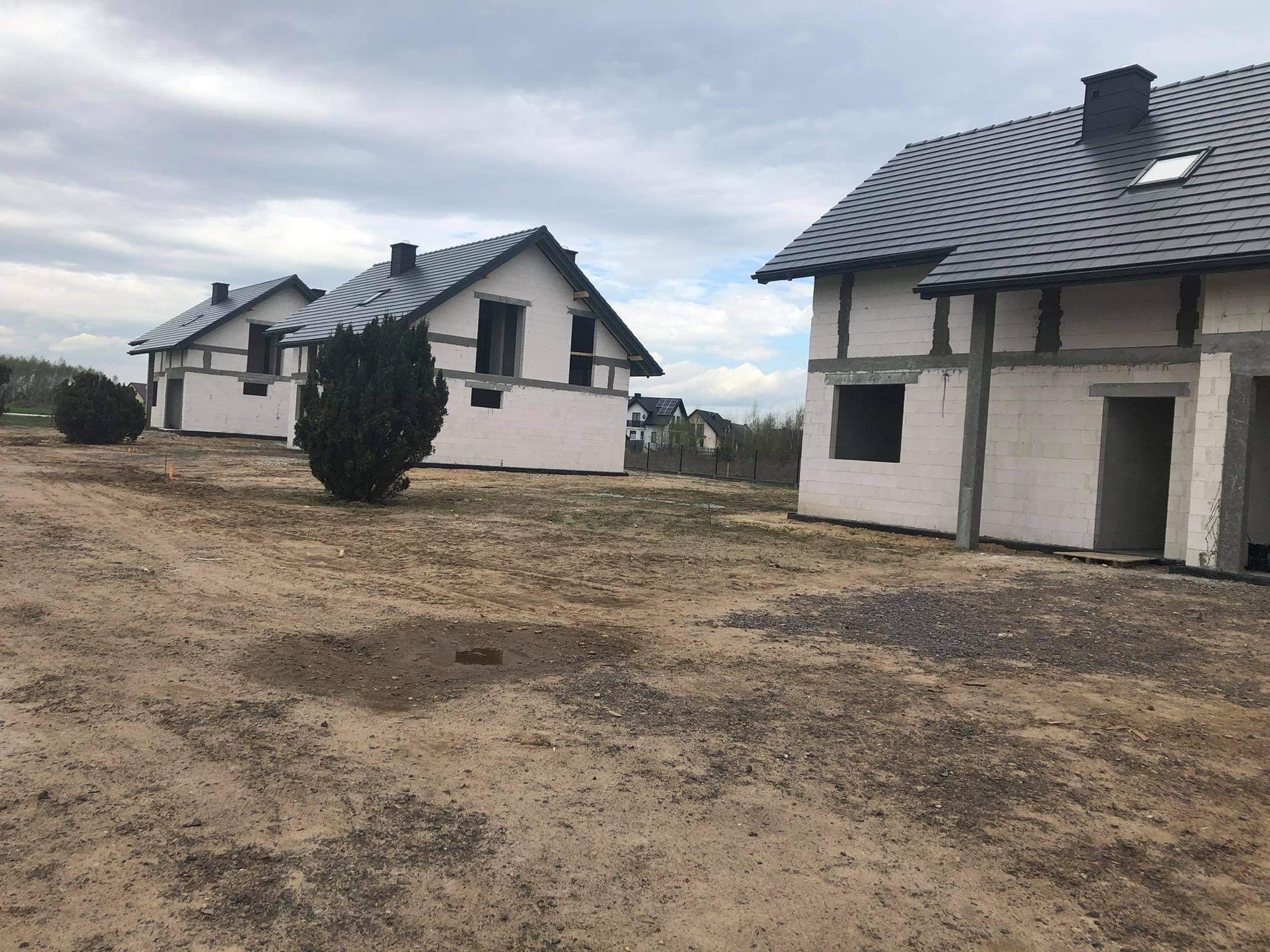 Trzy domy w trakcie budowy, wzniesione z białych bloczków, z ciemnymi dachami, widoczne niezadaszone otwory okienne i drzwiowe, na pierwszym planie piaszczyste podłoże.