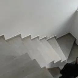 Schody wykończone jasnoszarymi płytkami, widok z góry, minimalistyczne wnętrze z białymi ścianami, w rogu schodów leży czarny przedmiot.