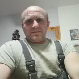 Portret mężczyzny w roboczym ubraniu na tle ściany z fotografiami i drewnianym zegarem z kukułką. Ujęcie z dołu,  oświetlenie mieszane.