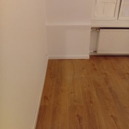 Tom Ltd - Jasne panele podłogowe w pokoju po remoncie, widok na ścianę pomalowaną na biało, grzejnik i okno w tle. Ujęcie z niskiej perspektywy.