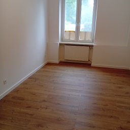 Tom Ltd - Puste, jasne wnętrze po remoncie: świeżo pomalowane ściany, nowe panele podłogowe w odcieniach brązu, białe okno z widokiem na zieleń. Minimalistyczny styl.