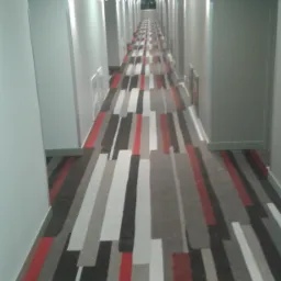 Długi korytarz hotelowy z geometrycznym wzorem w pasy w odcieniach szarości, czerwieni i czerni na podłodze, białe ściany z delikatnym oświetleniem punktowym, perspektywa zbieżna w kierunku okna...