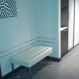 Tom Ltd - Świeżo pomalowane, błękitne ściany z zamontowanymi listwami przypodłogowymi oraz białą, pikowaną ławeczką na metalowych nogach, uzupełnione abstrakcyjnym obrazem w czarno-białe pasy.