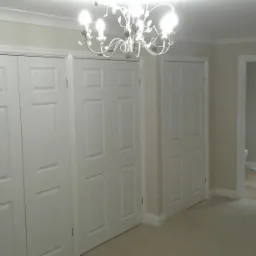 Jasne wnętrze po odświeżeniu: trzy białe, panelowe drzwi szafy wnękowej, kremowe ściany i sufit z listwami, w tle widoczne pomieszczenie z drewnianą podłogą i toaletą, oświetlone przez żyrandol.