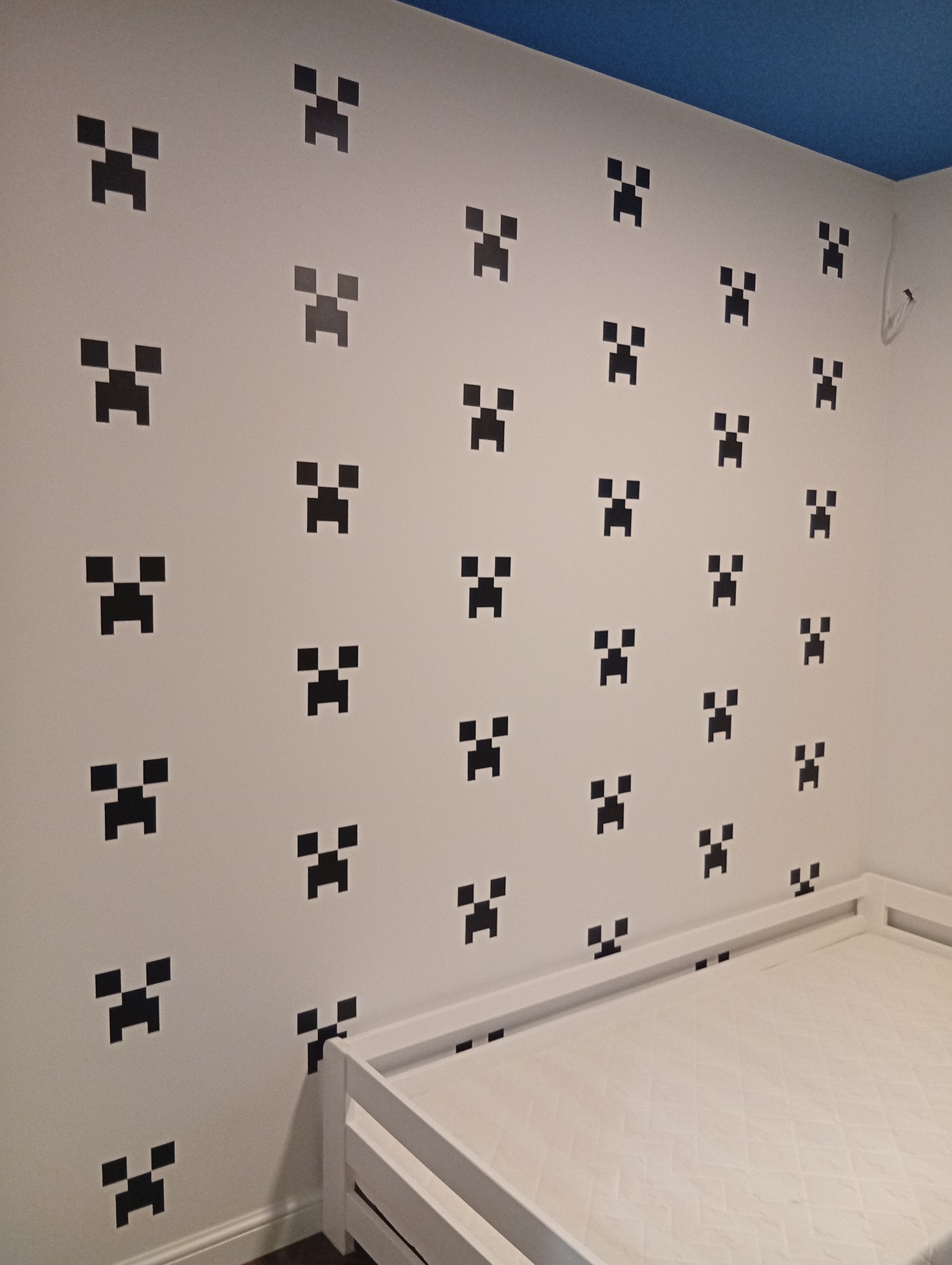 Pokój dziecięcy po dekoracji: biała ściana z czarnymi motywami Creeper z Minecrafta, niebieski sufit i białe łóżko. Nietypowy wzór, nowoczesny design.