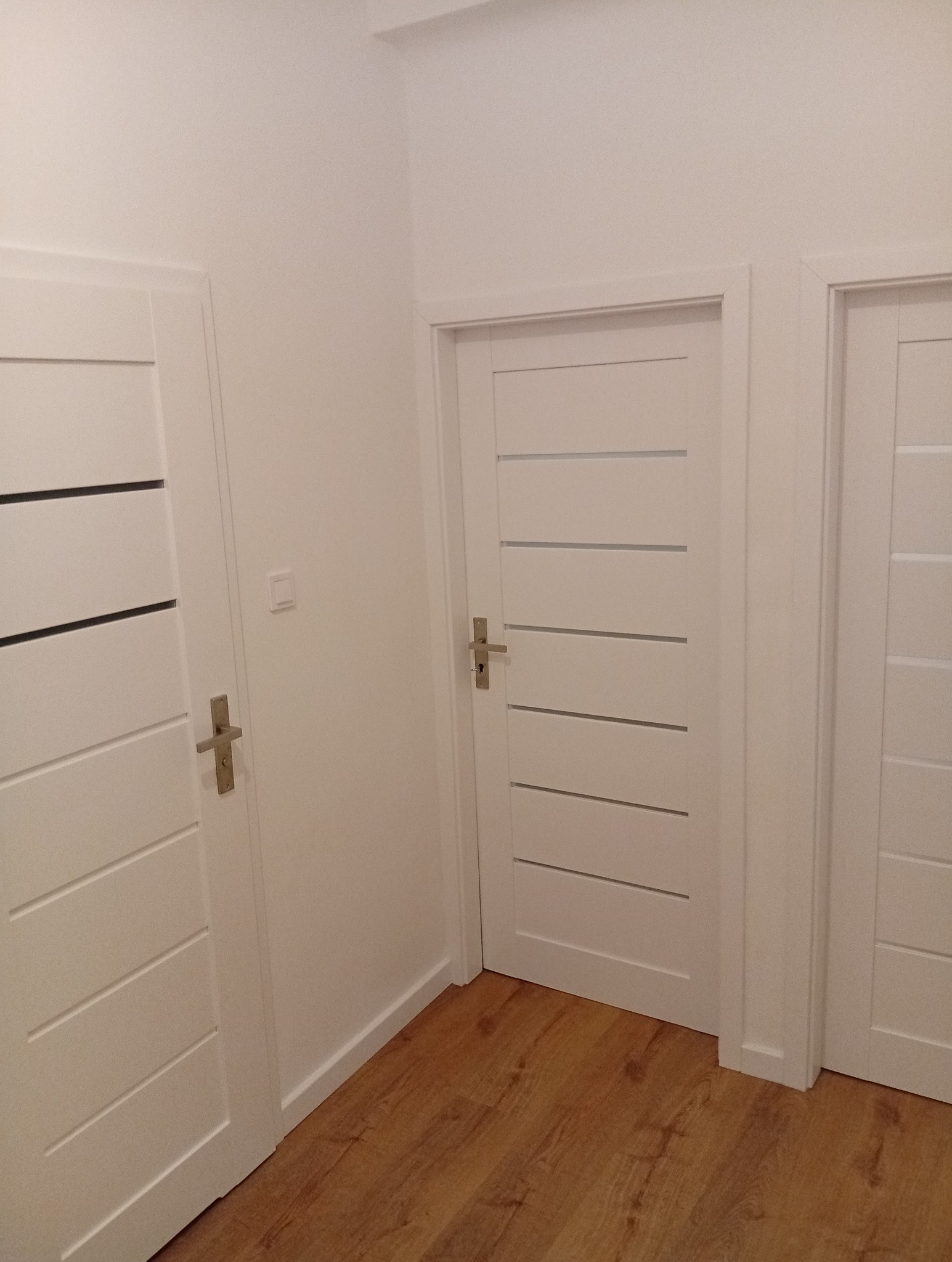 Jasne, minimalistyczne wnętrze po remoncie w Warszawie. Trzy białe drzwi z poziomymi frezami, drewnopodobna podłoga, świeżo pomalowane ściany.