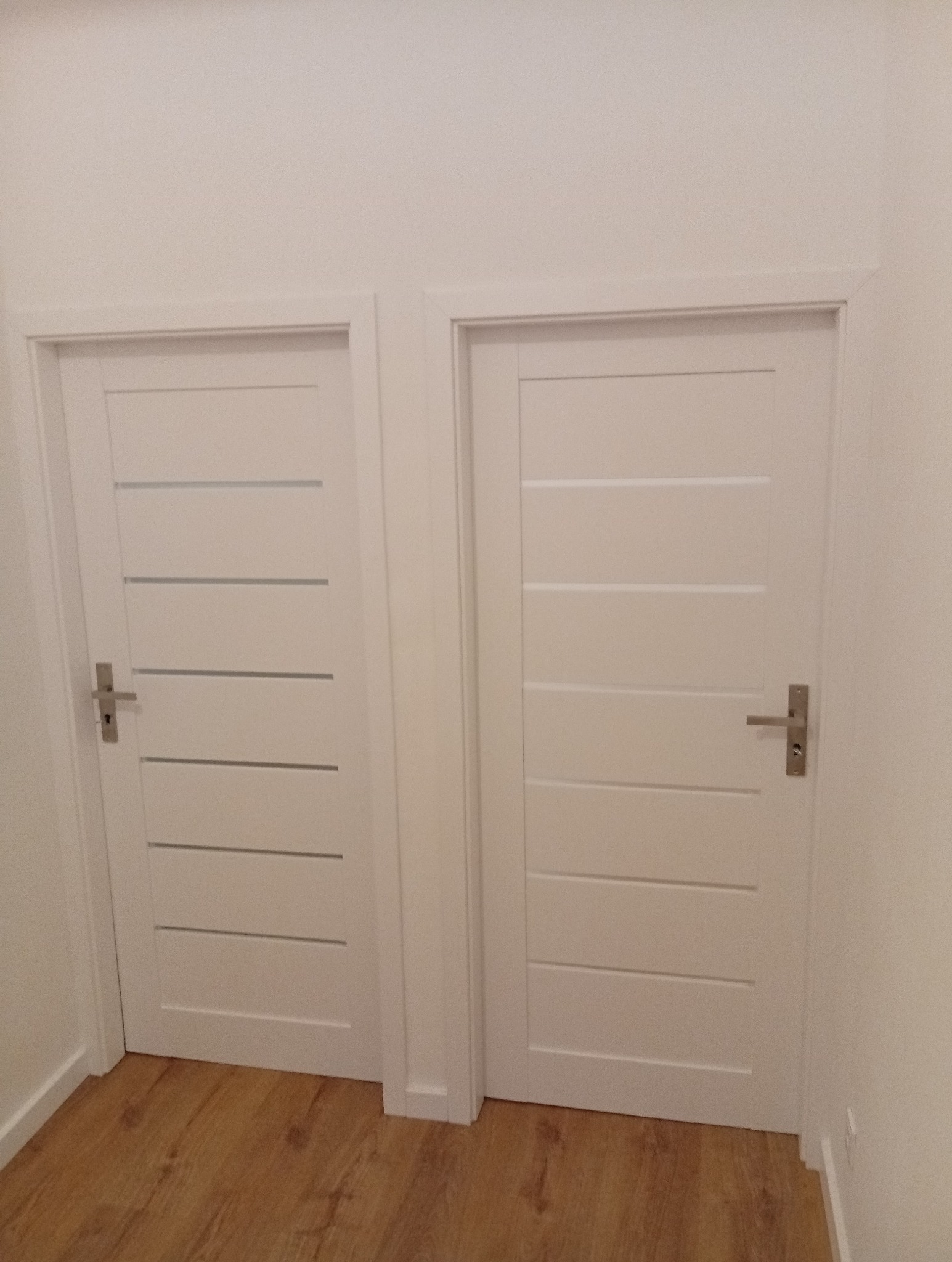 Dwa białe, identyczne drzwi wewnętrzne z poziomymi przeszkleniami i srebrnymi klamkami, zamontowane w białej ścianie nad panelami podłogowymi.