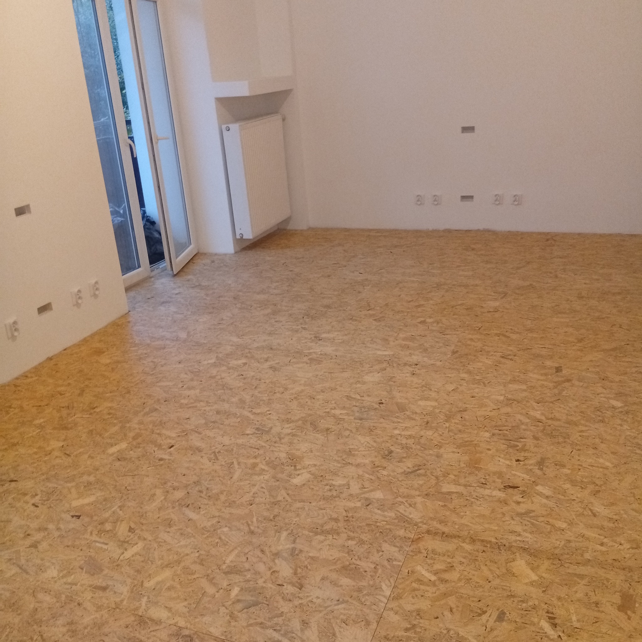 Surowa podłoga z płyt OSB w jasnym wnętrzu przed dalszymi pracami wykończeniowymi. Widoczne gniazdka elektryczne i grzejnik. Otwarty balkon w tle.