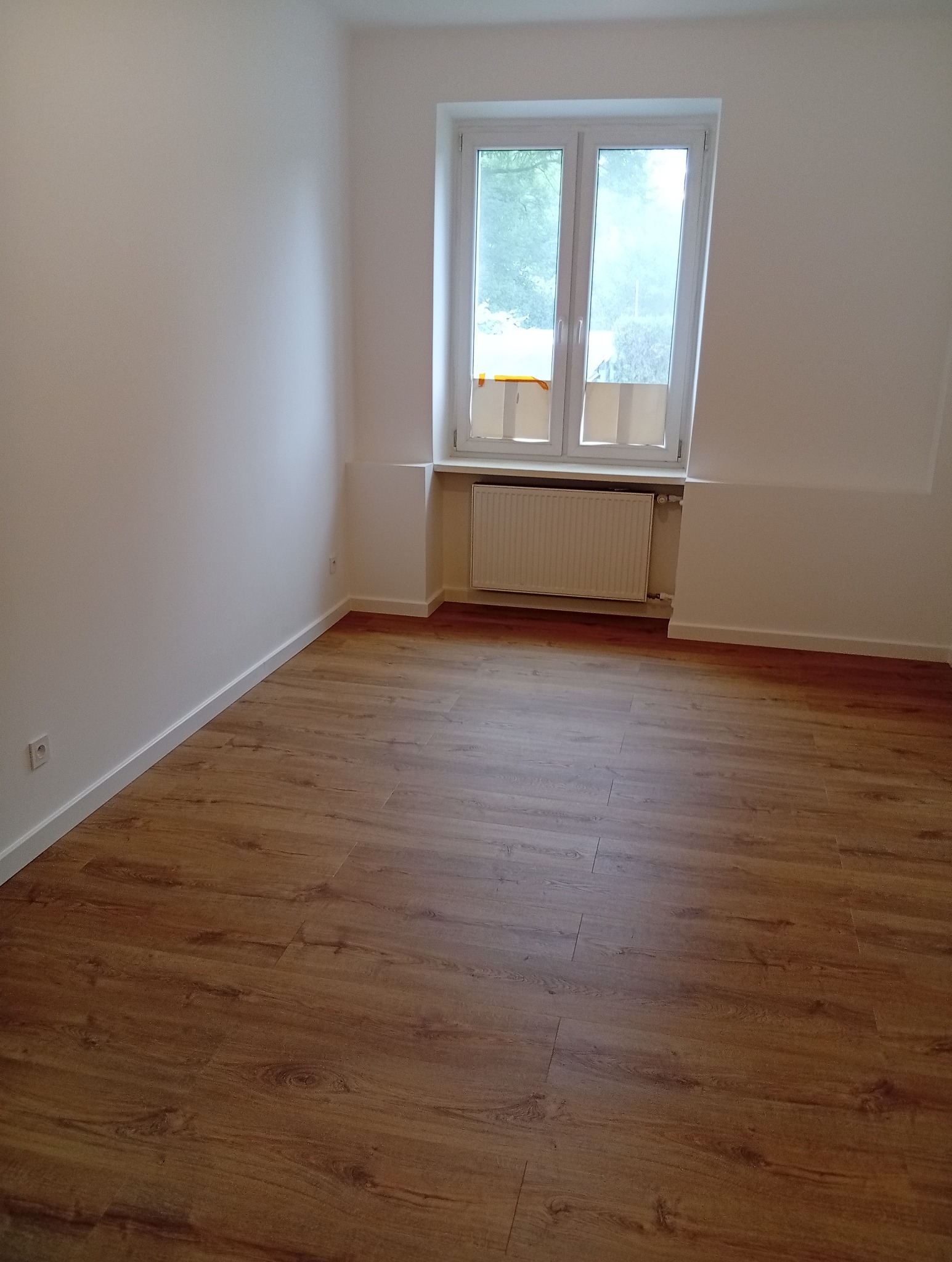 Puste, jasne wnętrze po remoncie: świeżo pomalowane ściany, nowe panele podłogowe w odcieniach brązu, białe okno z widokiem na zieleń. Minimalistyczny styl.