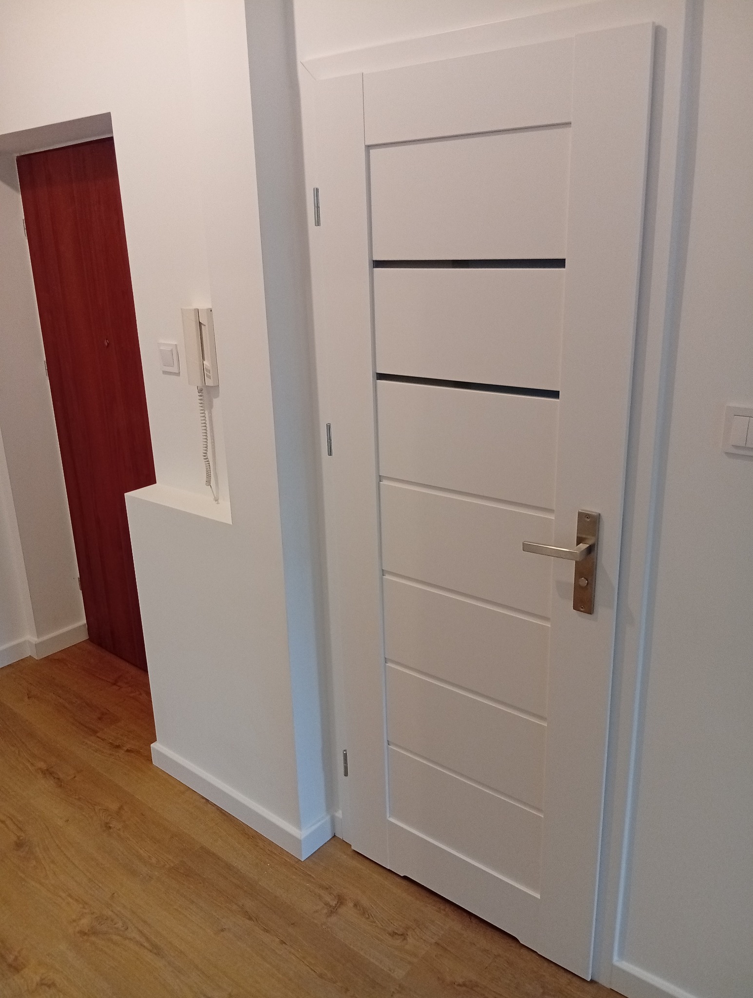 Białe, nowoczesne drzwi wewnętrzne z poziomymi frezami, zamontowane w odnowionym korytarzu z panelami podłogowymi i świeżo pomalowanymi ścianami na biało.