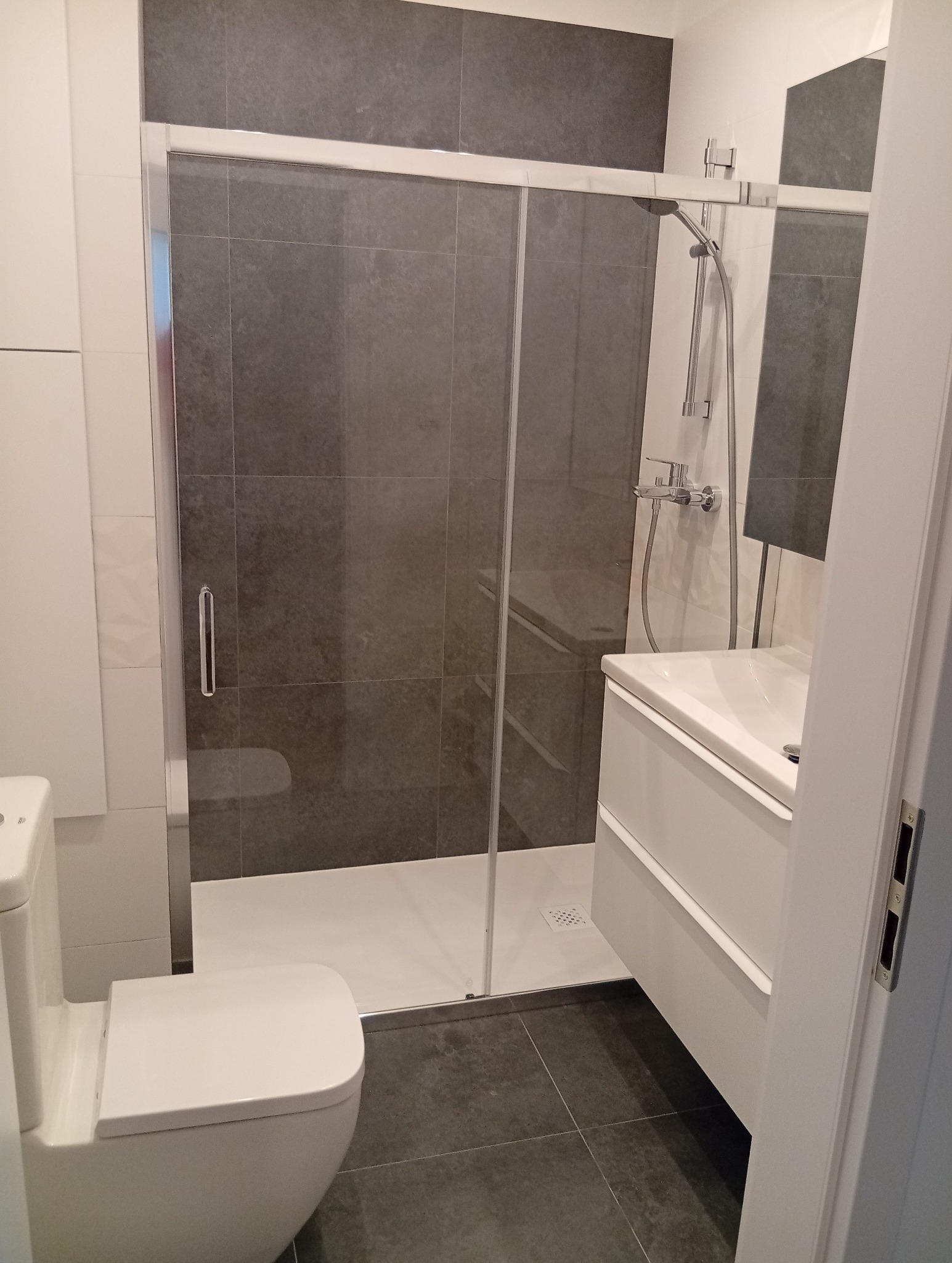 Nowoczesna łazienka po remoncie: kabina prysznicowa z szarymi płytkami, biała umywalka z szafką i toaleta. Minimalistyczny design, czyste linie, stonowana kolorystyka.