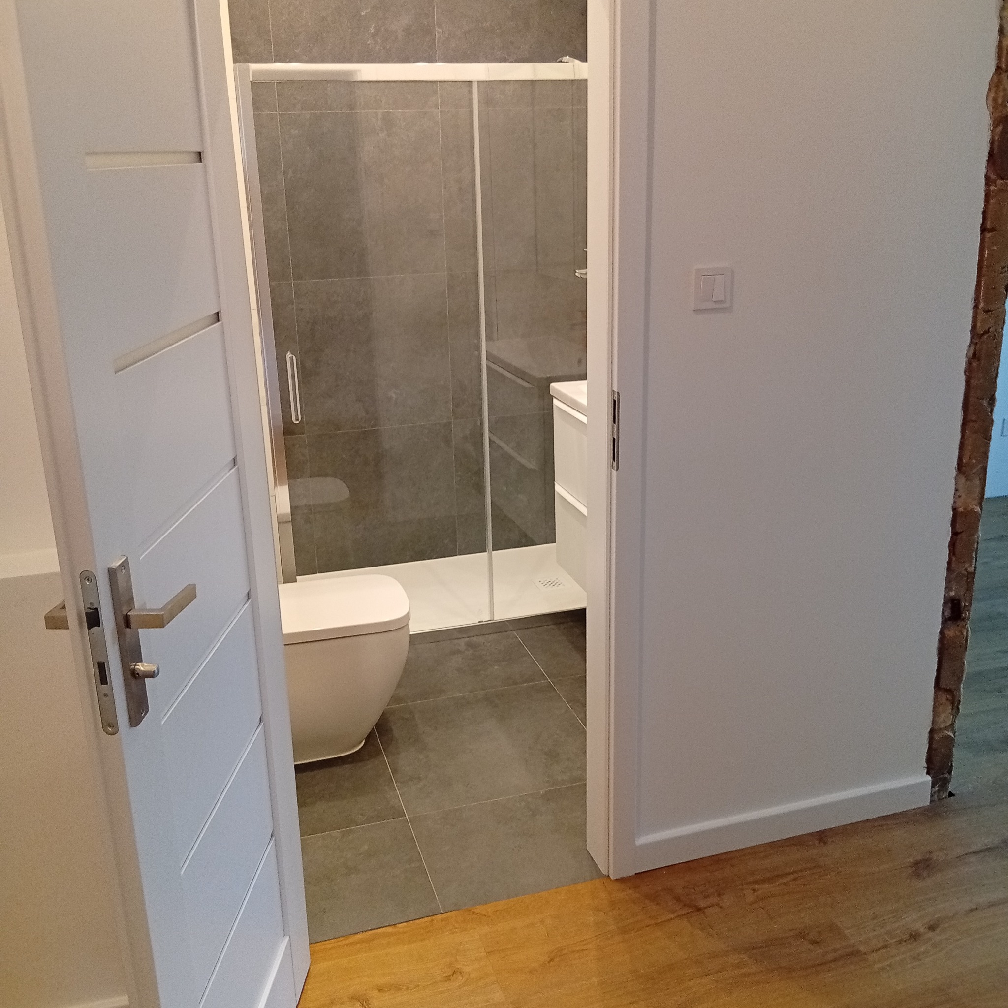 Wykończona łazienka z kabiną prysznicową i toaletą, widoczna przez otwarte białe drzwi z poziomymi frezami. Szare płytki na ścianach i podłodze, drewniana podłoga w przedpokoju.