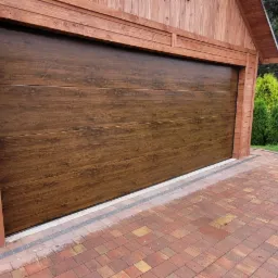 Zewnętrzny widok zamkniętej, segmentowej bramy garażowej w kolorze ciemnego drewna, zamontowanej w drewnianej konstrukcji garażu, z kostką brukową przed nią i żywopłotem w tle.
