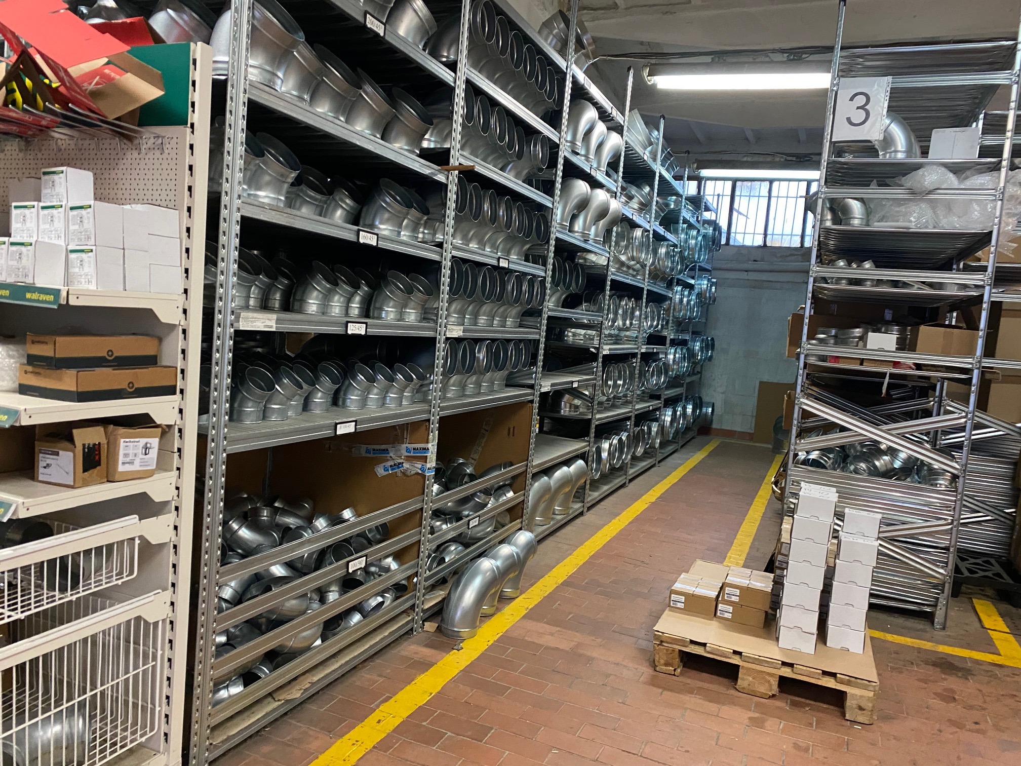 Magazyn z metalowymi kształtkami wentylacyjnymi na półkach regałów, widoczne skrzynki i paleta z zapakowanymi elementami.
