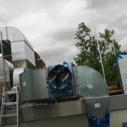 Instalacja wentylacyjna na dachu budynku, widoczne metalowe kanały wentylacyjne z wentylatorem, częściowo zabezpieczone folią, drabina i budynki w tle.