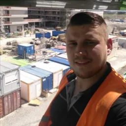 Uśmiechnięty mężczyzna w pomarańczowej kamizelce na budowie, w tle baraki i częściowo ukończony budynek.