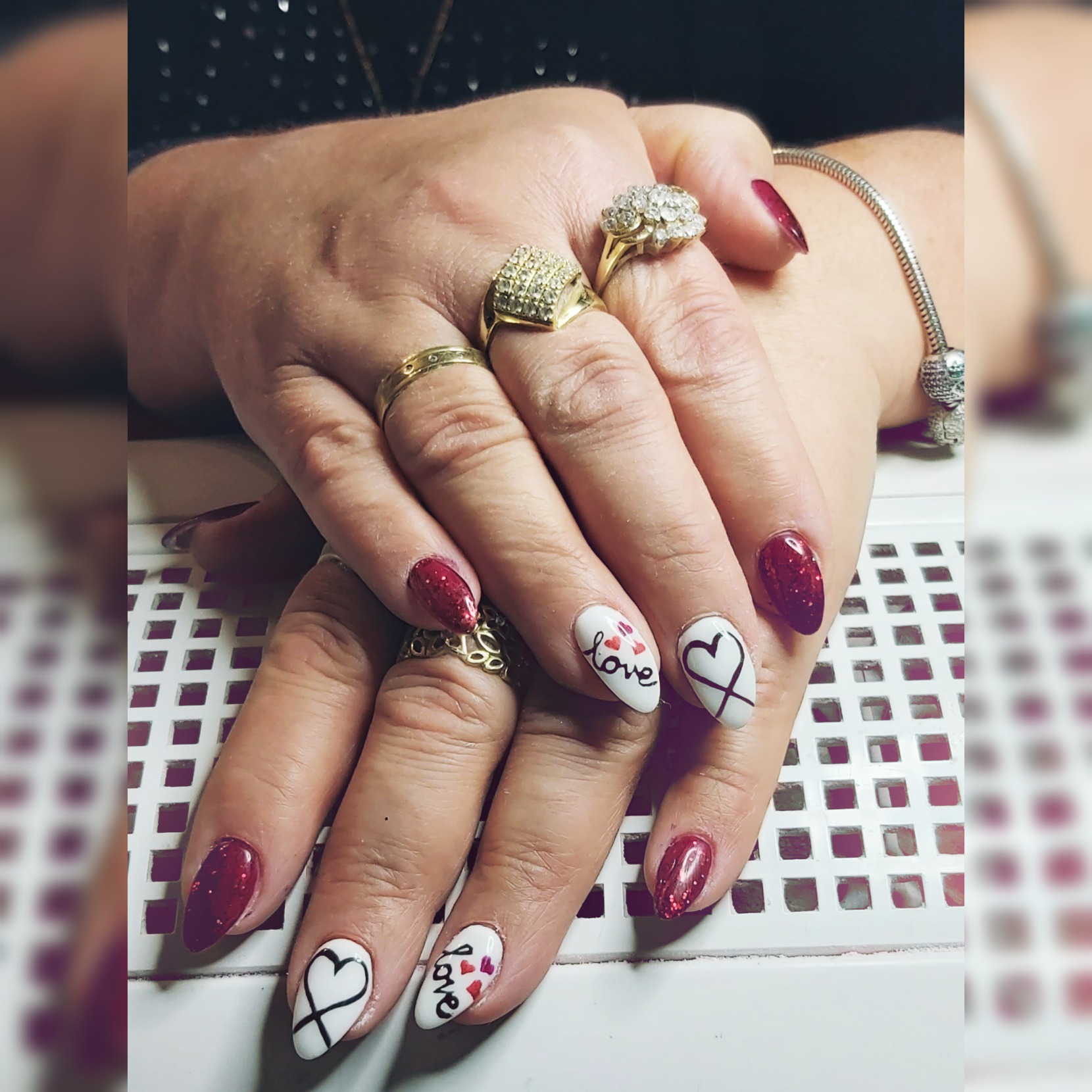 Dłonie kobiety z czerwono-białym manicurem walentynkowym: czerwone brokatowe paznokcie i białe z motywami serc i napisem 'love', kilka złotych pierścionków na palcach.