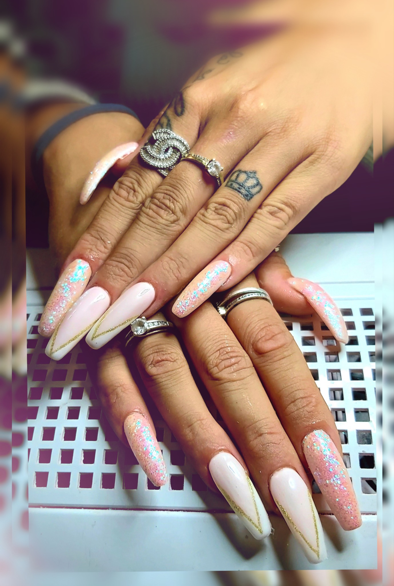 Dłonie z długimi, pomalowanymi paznokciami w odcieniach różu i bieli z brokatem i złotymi paskami, zdobione pierścionkami i tatuażem korony na palcu.