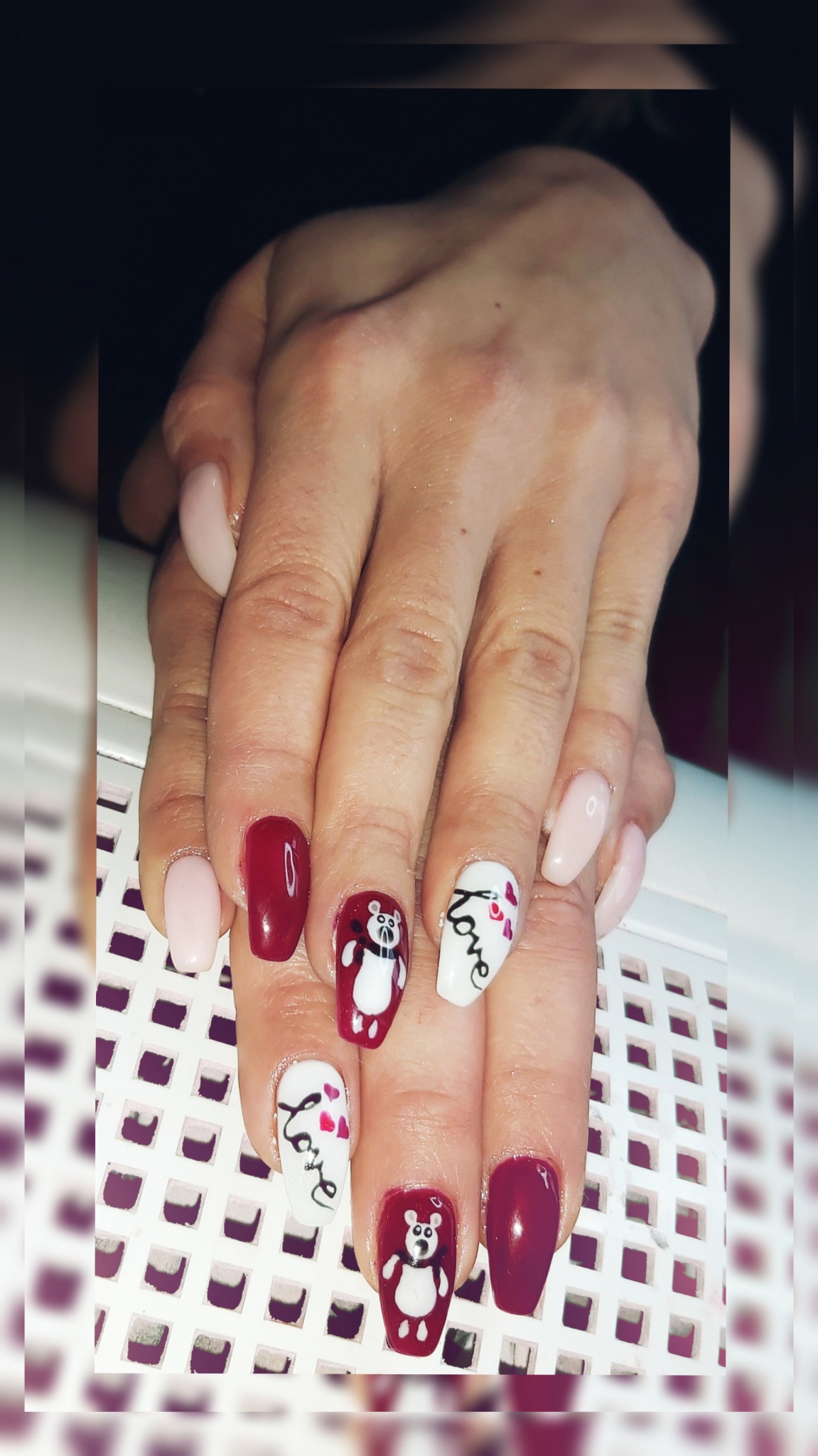 Dłonie z manicure hybrydowym: dwa paznokcie w kolorze burgundu, dwa w odcieniach nude, pozostałe z ręcznie malowanymi zdobieniami - misie i napisy 'love' na białym tle.