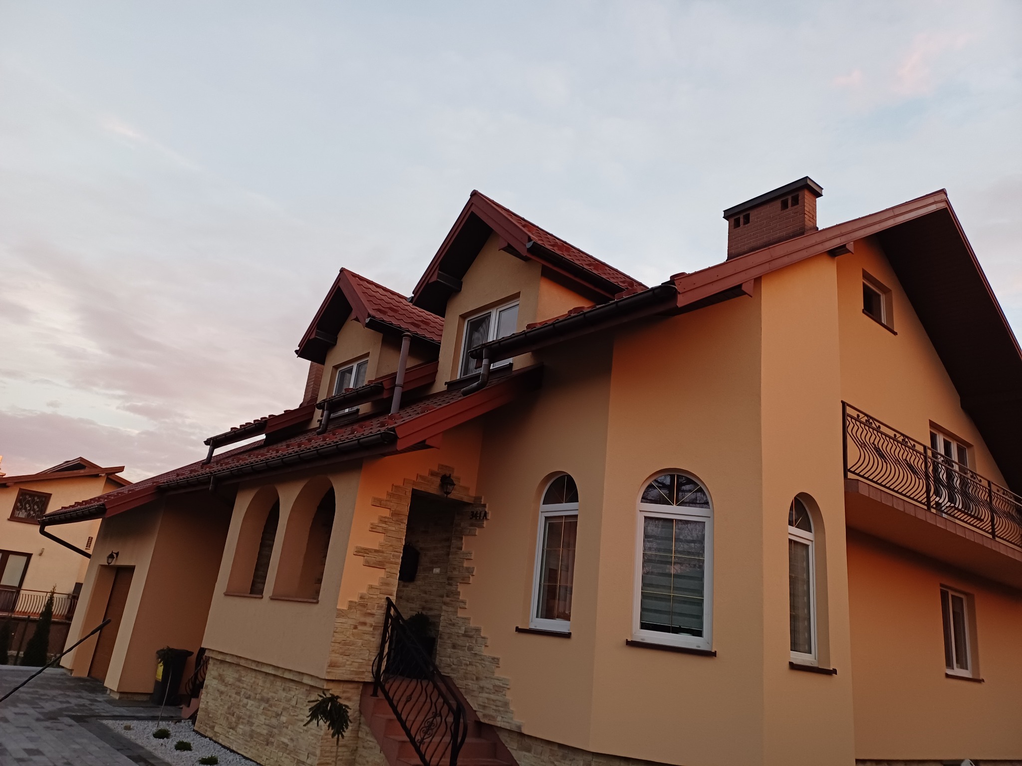 Elewacja domu jednorodzinnego z balkonem i schodami wejściowymi, ocieplona i pomalowana na beżowo, z elementami dekoracyjnymi z kamienia. Widok z dołu na tle nieba.