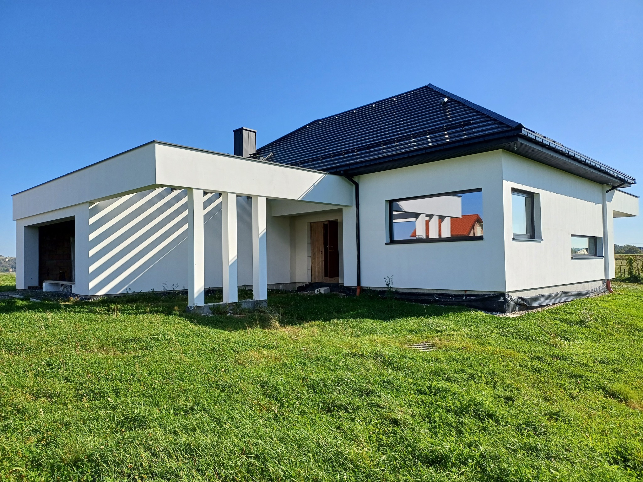 Nowoczesny, parterowy dom z płaskim dachem i garażem w bryle, wykończony na biało, z czarną dachówką, otoczony zielonym trawnikiem w słoneczny dzień.