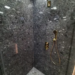 Kabina prysznicowa wykończona ciemnym, wzorzystym kamieniem z złotą armaturą: deszczownica, słuchawka prysznicowa z wężem oraz bateria.