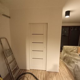 Tomasz kulig - Białe drzwi wewnętrzne z poziomymi frezami w świeżo pomalowanym pokoju. Widoczna drabina, odkurzacz i zabezpieczone folią meble. Podłoga wyłożona panelami.