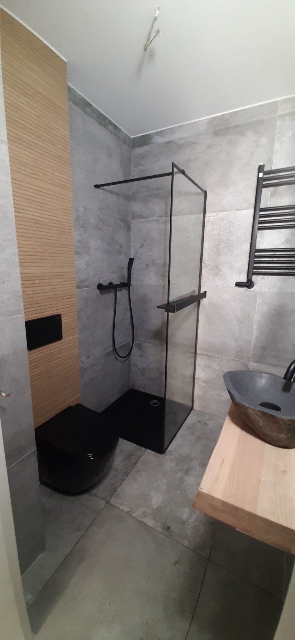 Nowoczesna łazienka z czarną armaturą, szklaną kabiną prysznicową typu walk-in, podwieszaną toaletą i umywalką w stylu kamiennym na drewnianym blacie, szare płytki imitujące beton na ścianach...