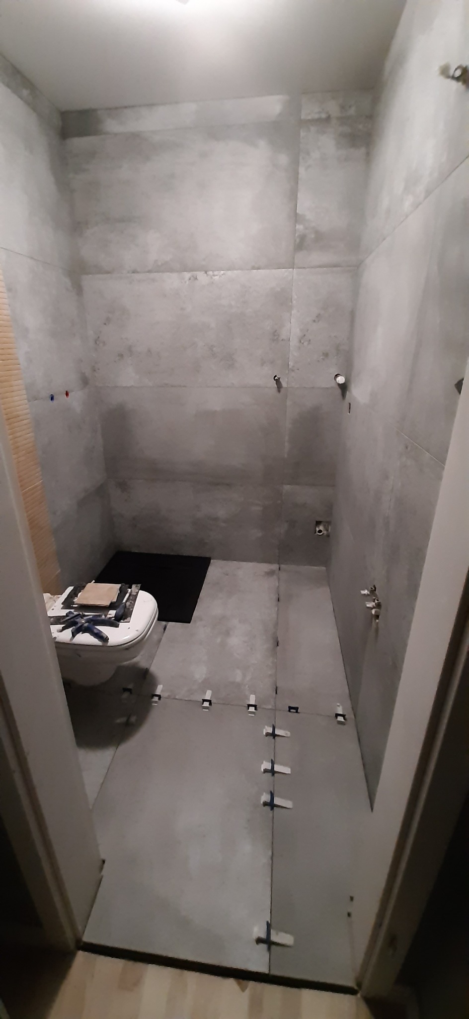 Łazienka w trakcie remontu: szare płytki na ścianach i podłodze, widoczne krzyżyki dystansowe, zamontowana toaleta z narzędziami na desce.