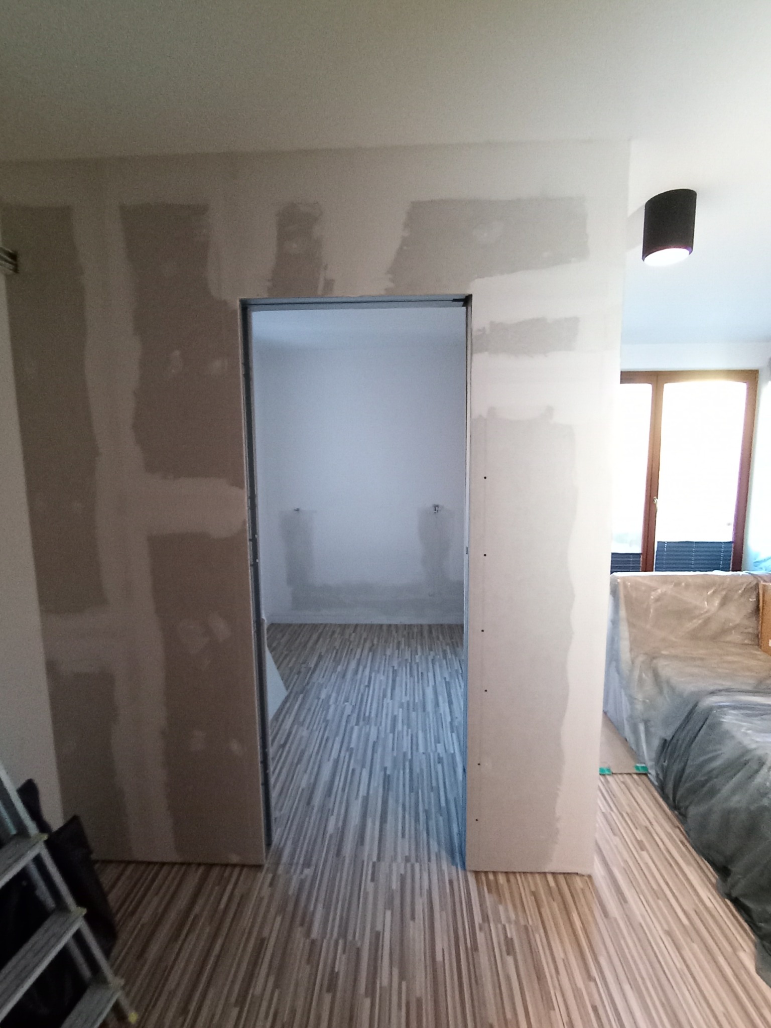 Wnętrze w trakcie prac wykończeniowych: ściany z płyt gipsowo-kartonowych z widocznymi łączeniami, podłoga wyłożona panelami, widoczny fragment drabiny i zakryty mebel.