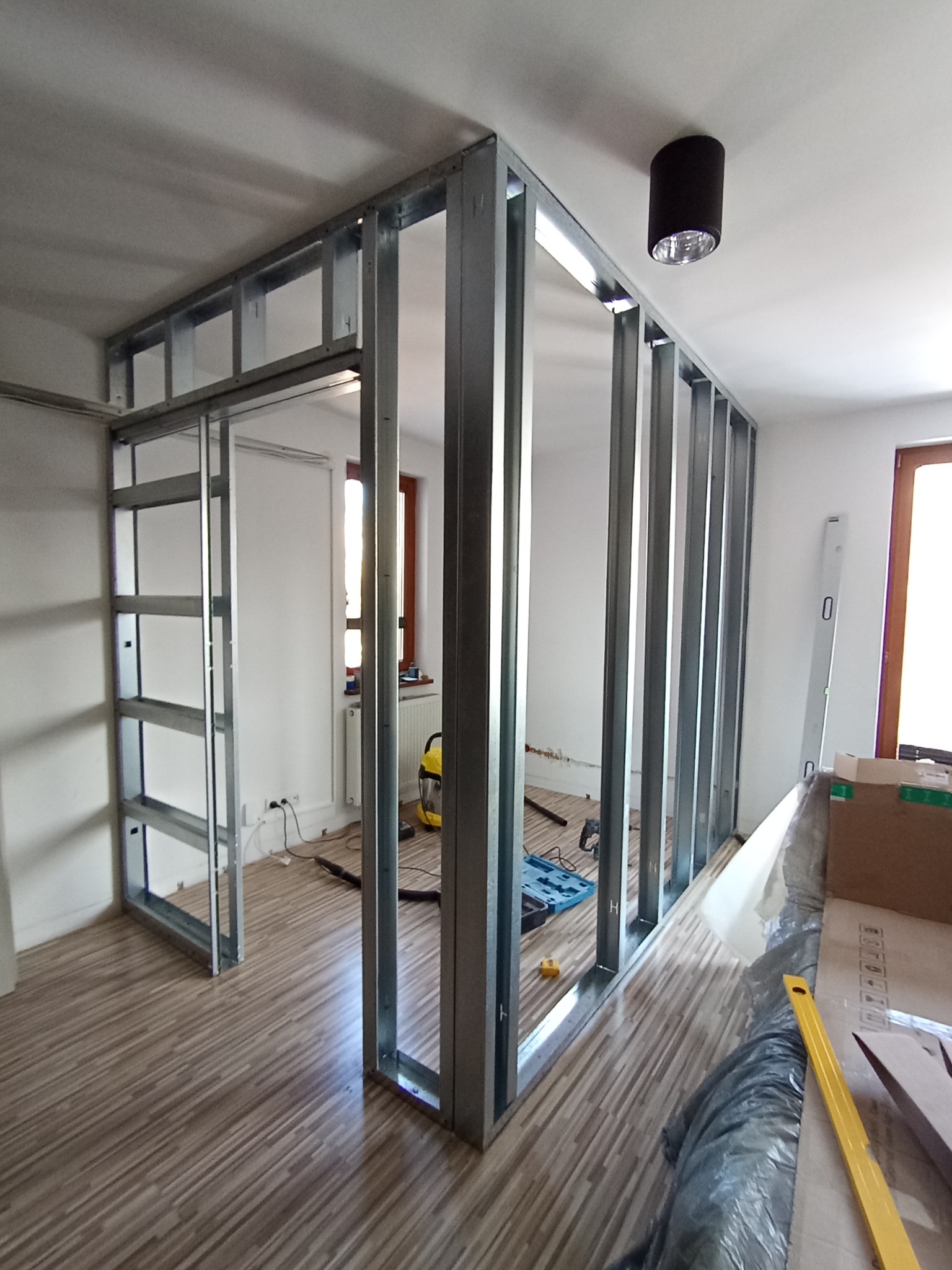 Montaż metalowej konstrukcji ścianki działowej w jasnym wnętrzu. Widoczne profile, narzędzia i fragmenty podłogi. Prace wykończeniowe w trakcie realizacji.