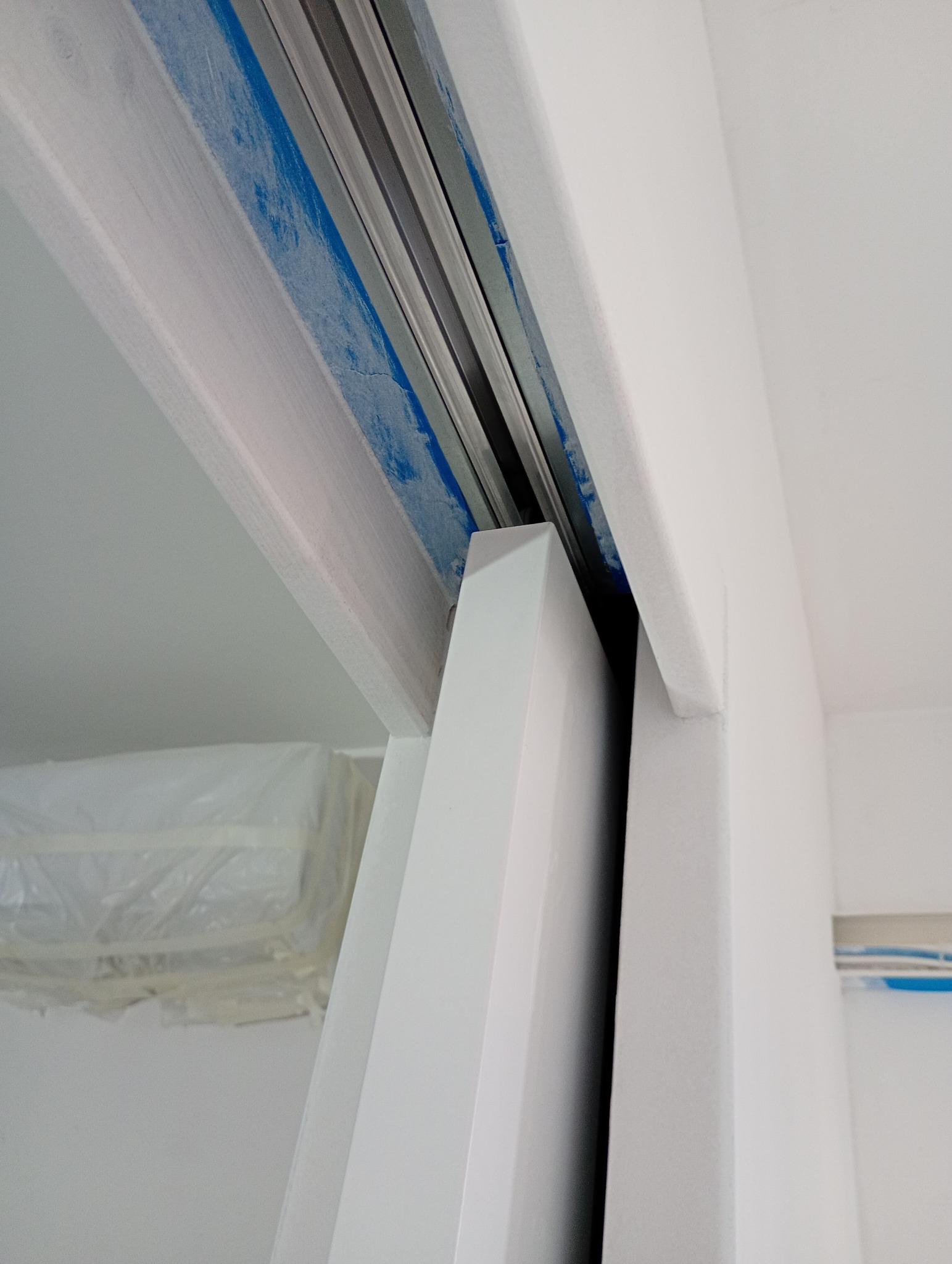 Białe, przesuwne drzwi w trakcie montażu, zabezpieczone niebieską taśmą malarską. Widoczna szyna i fragment ściany. Ujęcie z dołu.