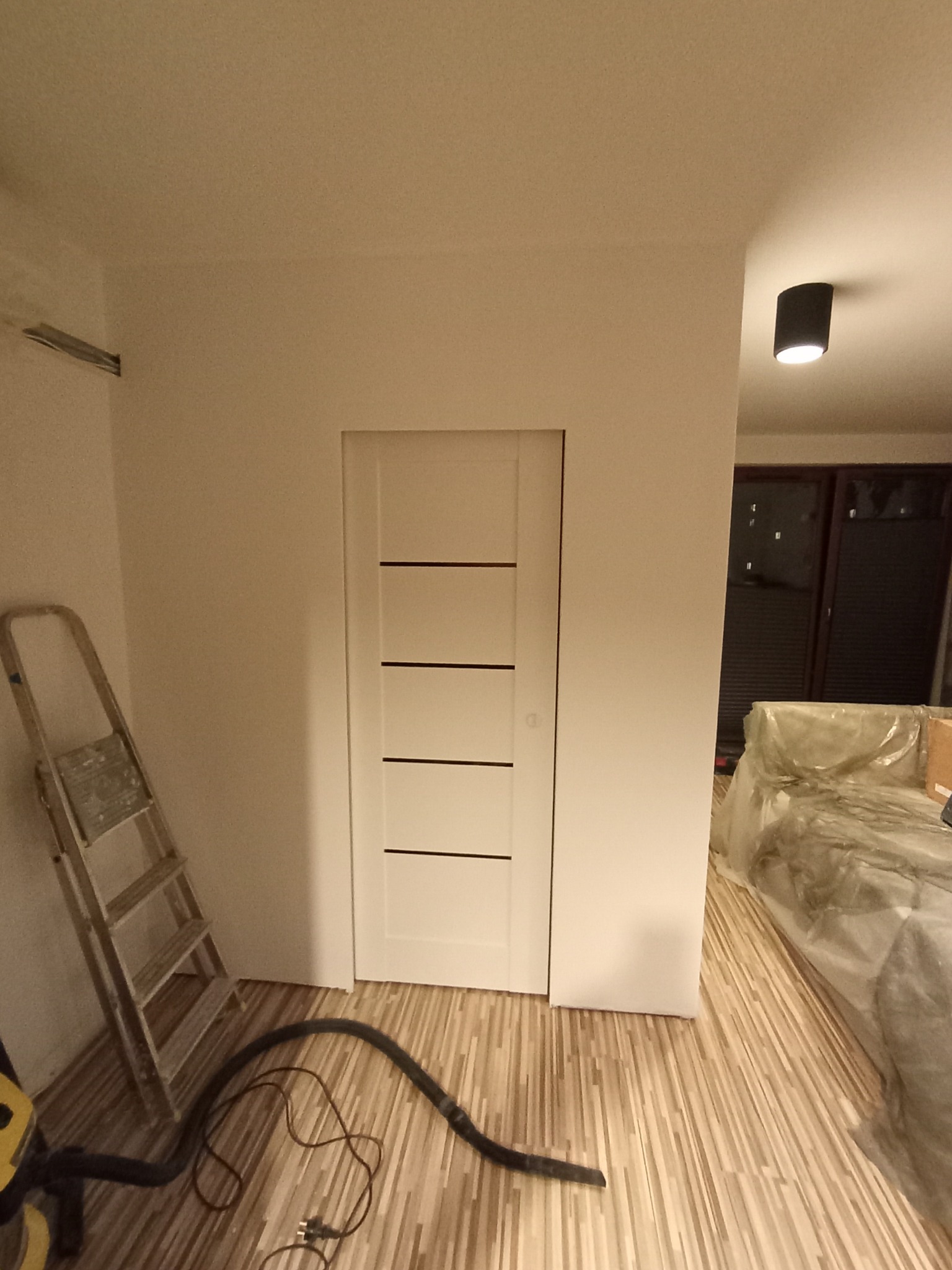 Białe drzwi wewnętrzne z poziomymi frezami w świeżo pomalowanym pokoju. Widoczna drabina, odkurzacz i zabezpieczone folią meble. Podłoga wyłożona panelami.