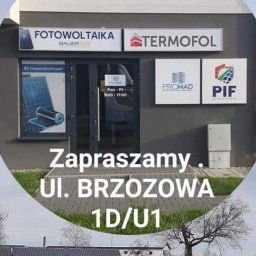 POLSKI INSTYTUT FOTOWOLTAIKI SPÓŁKA Z OGRANICZONĄ ODPOWIEDZIALNOŚCIĄ - Pracownik w czarnym ubraniu montuje panele na dachu budynku, w tle budynek firmy z szyldami Fotowoltaika, Termofol, Promad i PIF, widoczny adres Ul. Brzozowa 1D/U1.