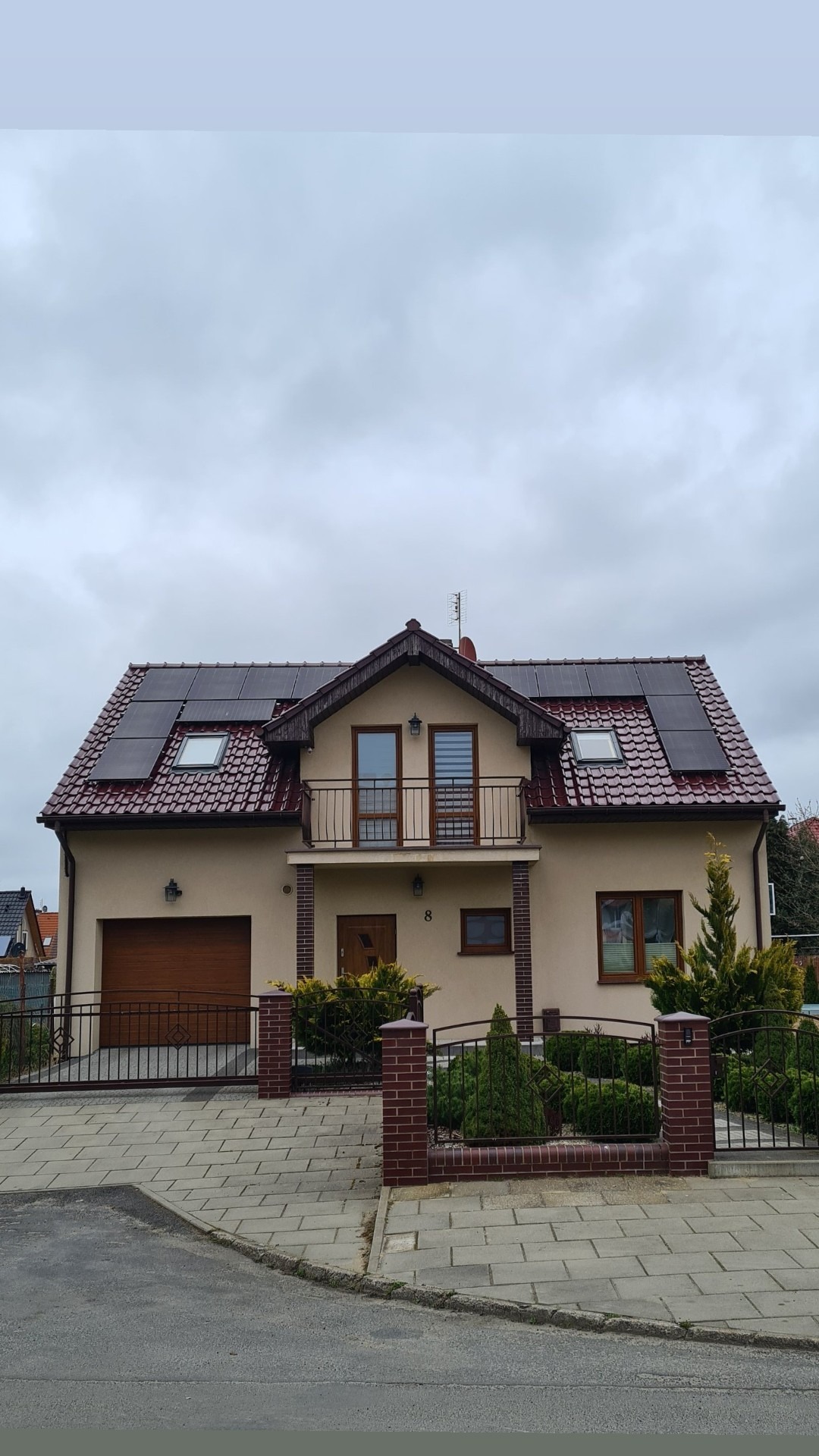 Dom jednorodzinny z zainstalowanymi modułami fotowoltaicznymi Bauer Glass Glass na dachu pokrytym czerwoną dachówką, widok z ulicy w pochmurny dzień.