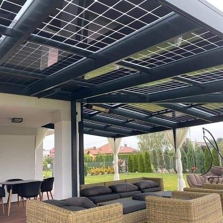Zadaszony taras z modułami fotowoltaicznymi w czarnej konstrukcji, widoczne meble ogrodowe z rattanu i białe zasłony, w tle domy i zieleń.