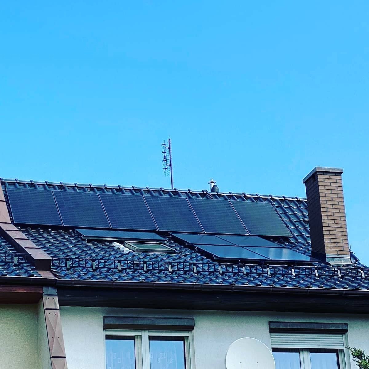 Panele słoneczne na ciemnym, skośnym dachu domu jednorodzinnego z brązową cegłą kominową i anteną telewizyjną na tle błękitnego nieba. Widoczne okno dachowe. Część paneli ma inny odcień.