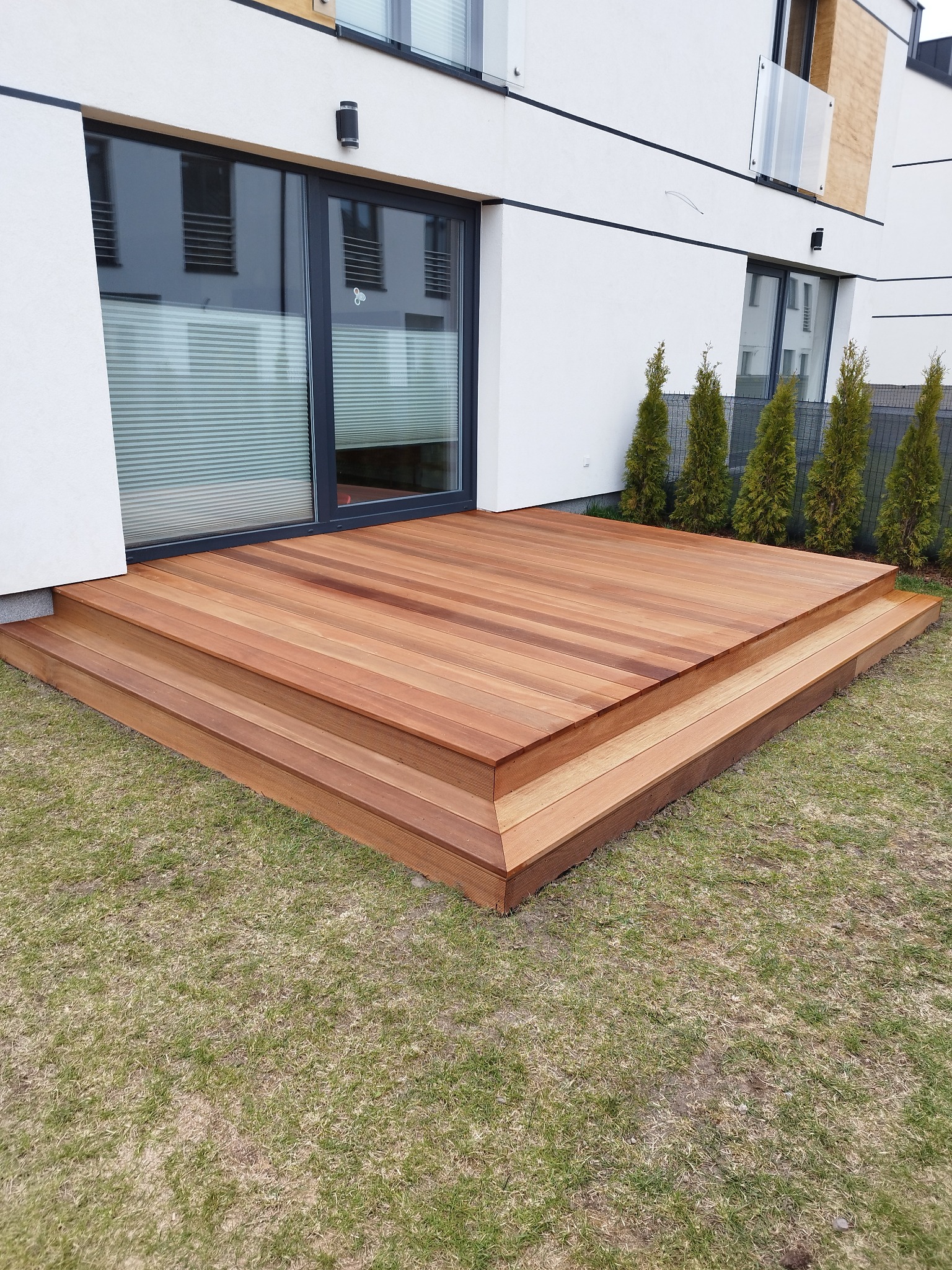 Dwupoziomowy taras z drewna egzotycznego, przylegający do nowoczesnego domu z dużym oknem tarasowym i rzędem małych drzew iglastych w tle.