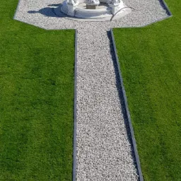 Widok z góry na ogród z fontanną otoczoną białymi kamieniami, ścieżką z jasnego żwiru obramowaną ciemnymi obrzeżami i równo przystrzyżonym trawnikiem, z przygotowanym miejscem pod nasadzenia w tle.