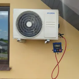 Zewnętrzna jednostka klimatyzacji Hyundai zamontowana na elewacji budynku, podłączona do cyfrowego kolektora pomiarowego z czerwonymi i żółtymi przewodami, okno w tle.