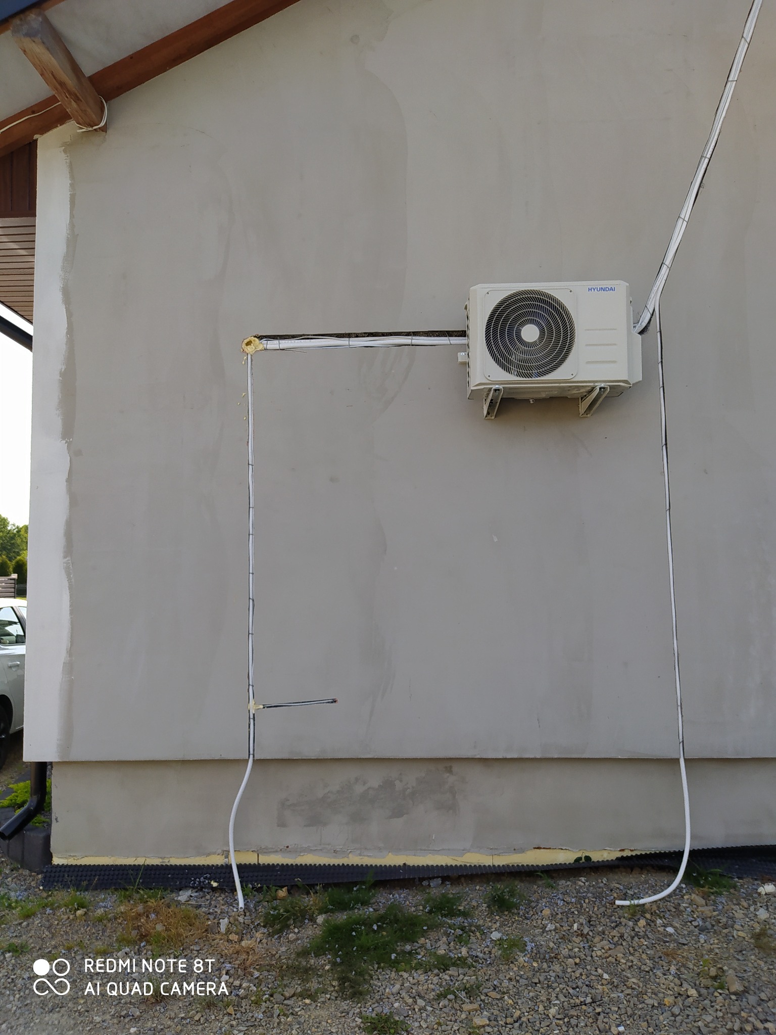 Zamontowana jednostka zewnętrzna klimatyzatora Hyundai na szarej elewacji budynku, widoczne rurki instalacyjne w białych osłonach, fragment dachu i otoczenie budynku z kamieniami.