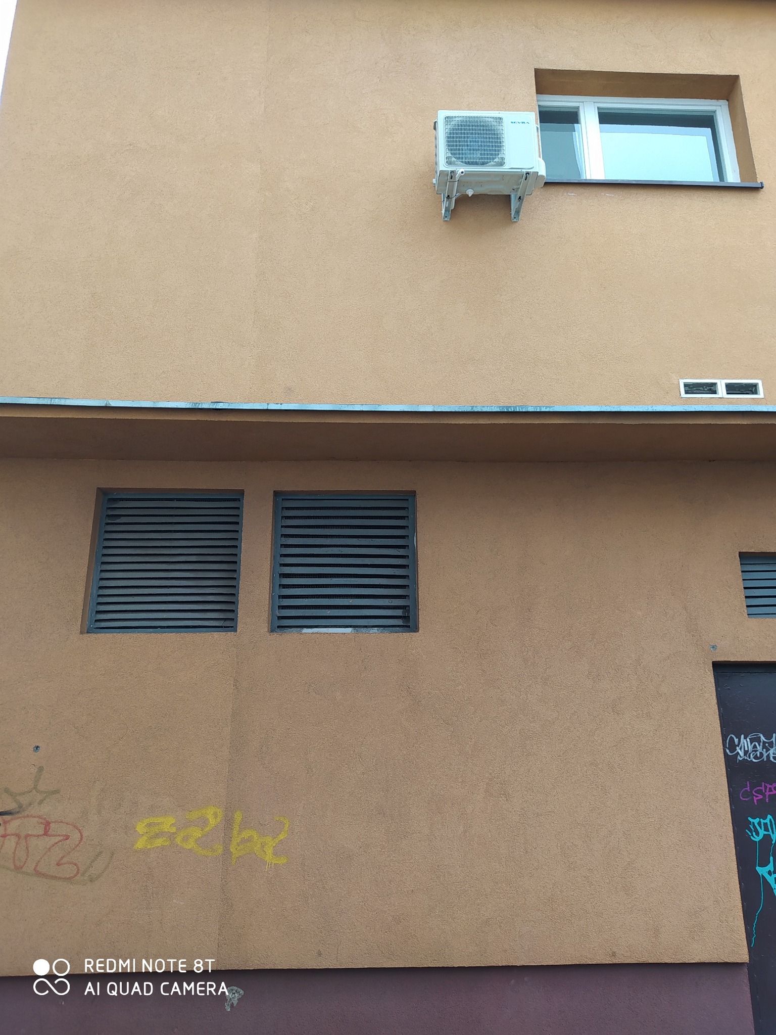 Zewnętrzna jednostka klimatyzacji zamontowana na ścianie budynku z oknem w tle, widoczne kratki wentylacyjne i graffiti na elewacji.