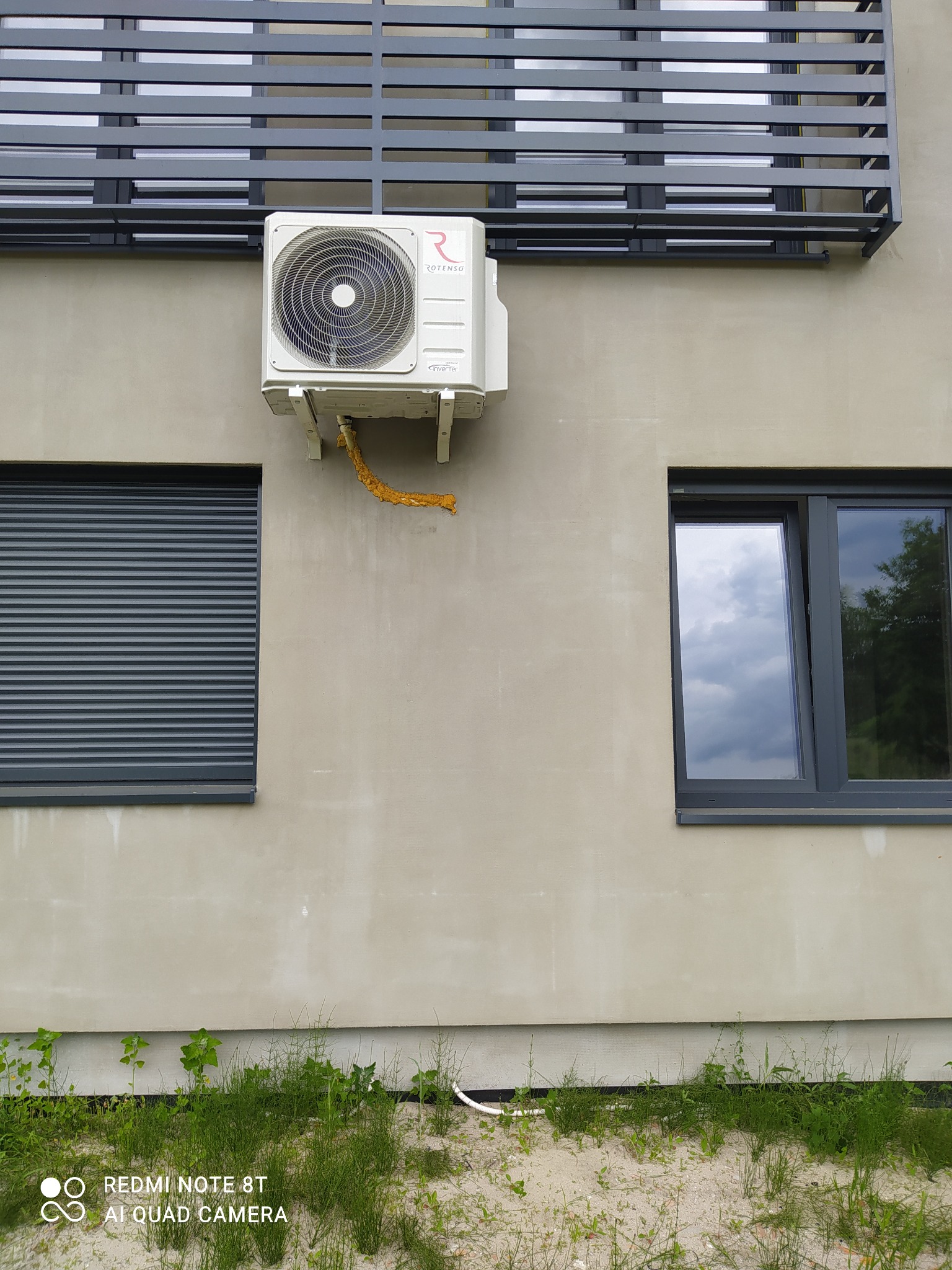 Zamontowany na elewacji budynku klimatyzator Rotenso z widoczną rurą odprowadzającą skropliny zabezpieczoną żółtą izolacją, poniżej okno z żaluzjami zewnętrznymi i trawa.