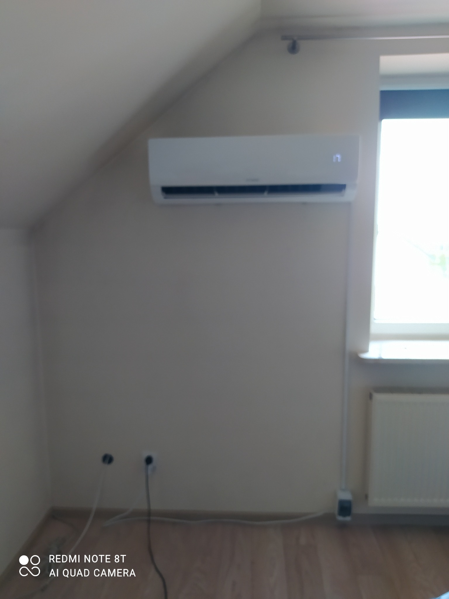 Biały klimatyzator na ścianie pod skosem na poddaszu, widoczne kable i otwory w ścianie przed podłączeniem.