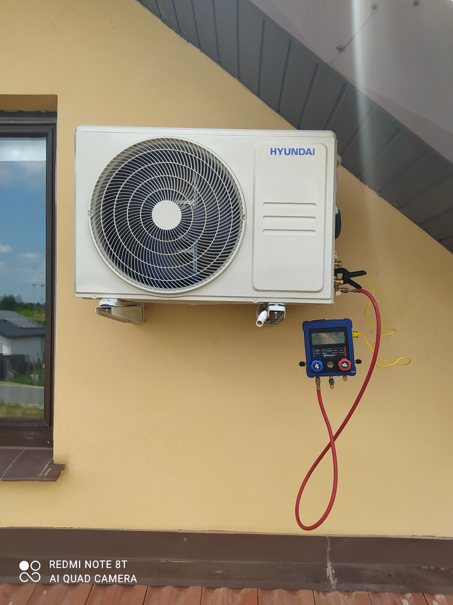 Zewnętrzna jednostka klimatyzacji Hyundai zamontowana na elewacji budynku, podłączona do cyfrowego kolektora pomiarowego z czerwonymi i żółtymi przewodami, okno w tle.